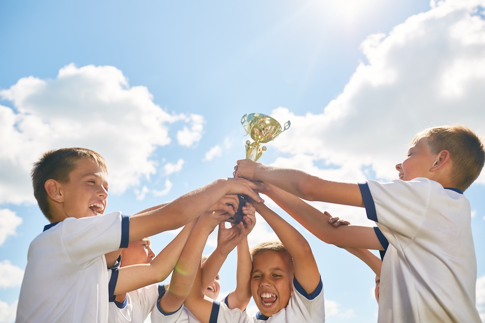 Team of children celebrating victory in sports, cheering and holding golden cup against clear blue sky and getting excited about local sports club sponsorship.